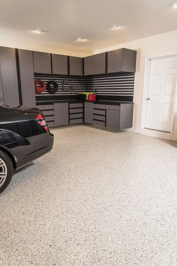 Light beige and brown flake garage floor with gray cabinets and a slatwall, and a black car at the left