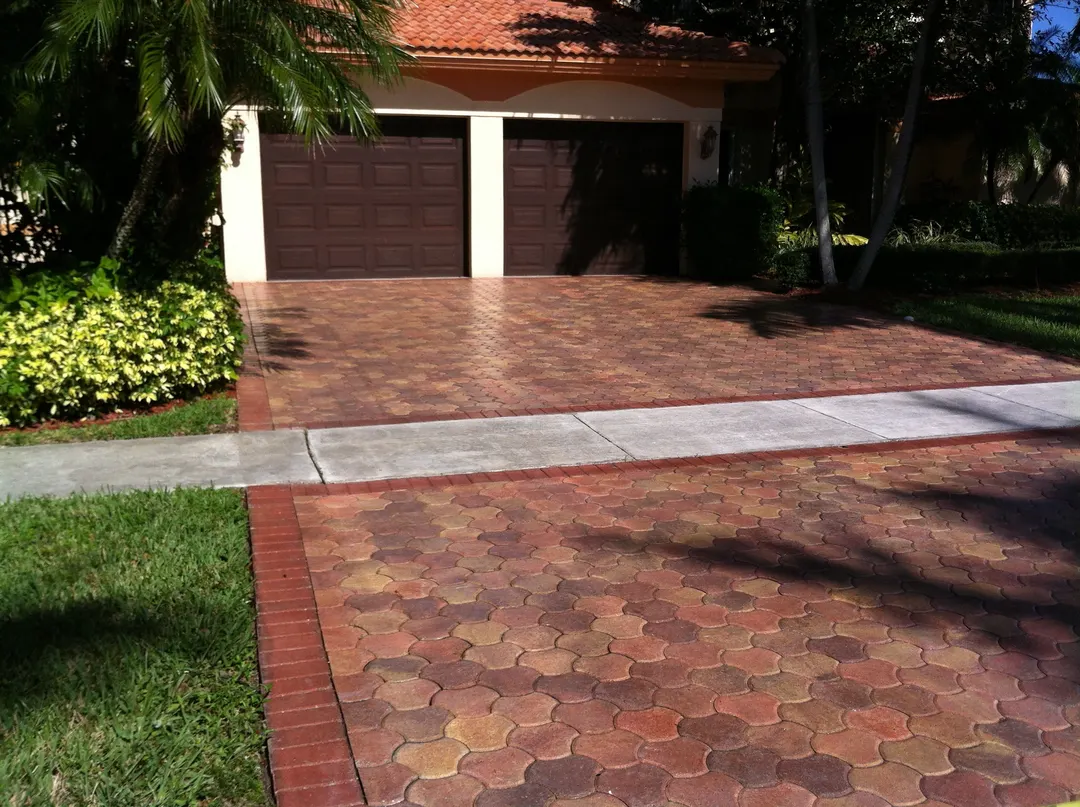Clay-colored paver driveway with a brick border leading to a two-car garage, surrounded by palms and shrubs